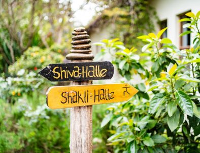 Wegweiser aus Holz mit den Aufschriften ‚Shiva-Halle‘ und ‚Shakti-Halle‘ vor grünem Gartenhintergrund in der Lykia Lodge.