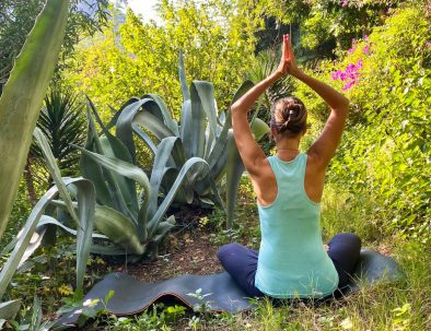 Frau praktiziert Yoga im Schneidersitz inmitten üppiger Natur, mit erhobenen Armen vor einer großen Agave.